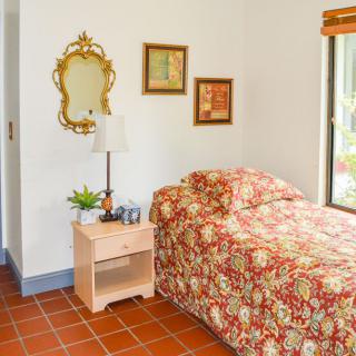 A cozy bedroom with a single bed featuring floral bedding, a wooden nightstand with a lamp and potted plant, and decorative wall art including a mirror.