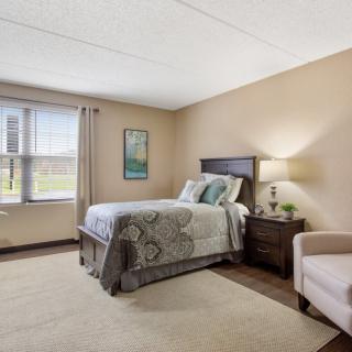 A cozy bedroom with a single bed, a nightstand with a lamp, and a comfortable chair by the window.