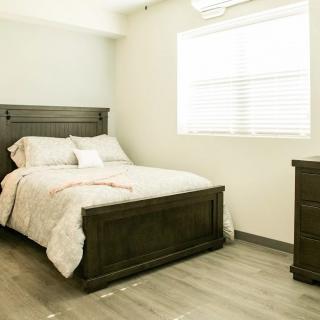 A minimalist bedroom with a wooden bed and dresser, featuring light walls and a large window.