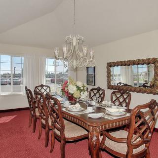 A formal dining room with a wooden table set for dinner, surrounded by ornate chairs and illuminated by a chandelier. The room features a red carpet and large windows, with a decorative mirror on the wall.