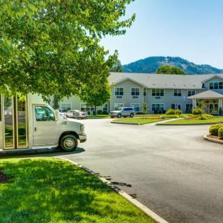 A sunny day with a white vehicle parked near a building surrounded by greenery and mountains.