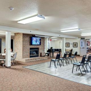 A well-lit meeting room with chairs arranged facing a television and fireplace, featuring a piano to the side.