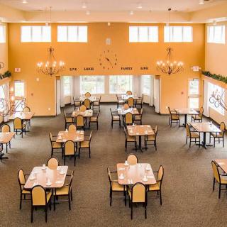 An elegant dining room with neatly arranged tables and chairs, lit by chandeliers and natural light from large windows.