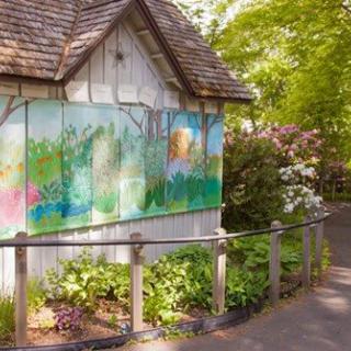 A quaint cabin with a colorful mural sits beside a lush garden and pathway under vibrant green trees.