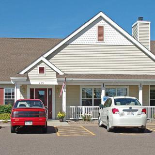 A small house with a front porch and two cars parked in the driveway on a sunny day.