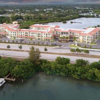 Aerial view of a large building complex near a body of water, surrounded by greenery and a road.