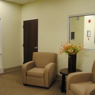 A waiting room with two beige chairs, a small table with flowers, and a door.