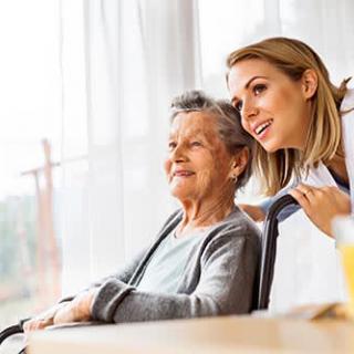 A caregiver smiling alongside an elderly woman in a wheelchair by the window.