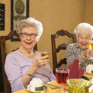 Two elderly women smiling and toasting with drinks at a dining table.
