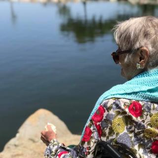 An elderly woman wearing sunglasses and a floral shawl, sitting by a lake enjoying the outdoors.