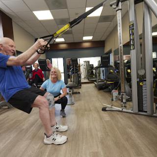 An older adult engages in a TRX workout, supported by a trainer in a vibrant gym environment.