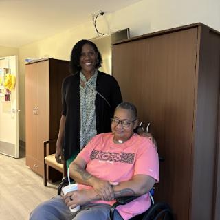 A caregiver stands beside a person in a wheelchair, both smiling in a comfortable indoor setting.