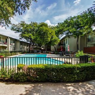 A serene view of an apartment complex featuring a fenced swimming pool surrounded by lush greenery on a sunny day.