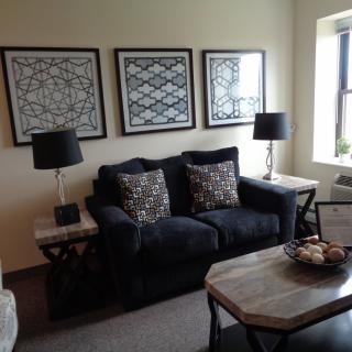 A cozy living room with a dark sofa, decorative cushions, and framed artwork above it. The space is complemented by side tables with lamps and a central coffee table.