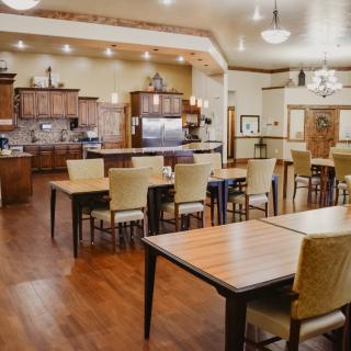 A spacious and well-lit dining area with a kitchen, featuring wooden tables and chairs on a polished wooden floor.