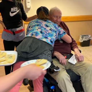 An elderly man in a wheelchair receives a hug from a caregiver while another person holds plates of food.