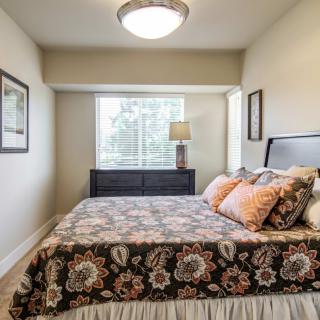 A cozy bedroom featuring a bed with floral bedding, a lamp on the dresser, and natural light streaming through the window.