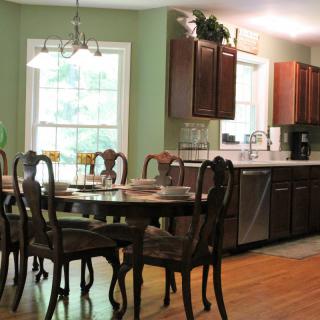 A cozy dining area with a wooden table and chairs, adjacent to a kitchen with dark cabinets and a large window.