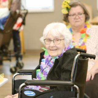 A joyful moment captured at a care facility, where an elderly woman in a wheelchair enjoys a lively gathering adorned with colorful flower leis.