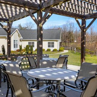 A sunlit patio area with tables and chairs under a pergola, overlooking a landscaped garden and building in the background.