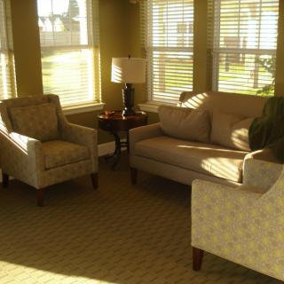 A cozy living room with natural light streaming through the windows, featuring a sofa, armchairs, and a lamp.