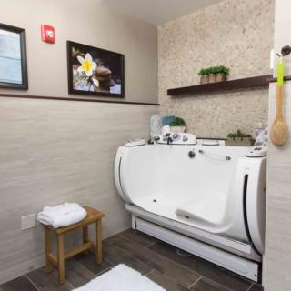 A modern bathroom featuring a spa-like bathtub with towels on a small wooden stool and decorative elements on the wall and shelf.