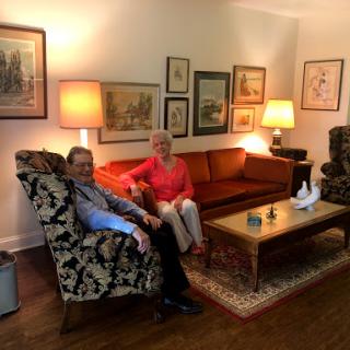 A cozy living room featuring a couple seated together on a patterned armchair and sofa, surrounded by art and warm lighting.
