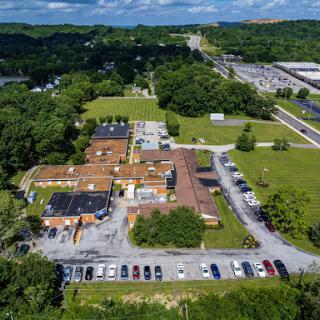 Aerial view of a building complex surrounded by greenery and a parking lot, with a roadway visible in the distance.