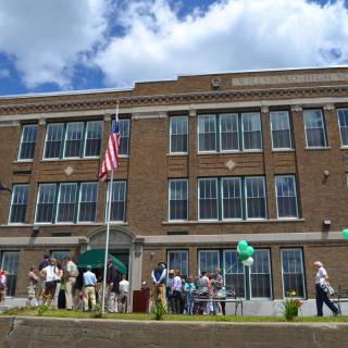 A group of people gathered outside a large brick building on a sunny day, with an American flag and green balloons visible.