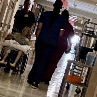 Medical staff assist a patient in a wheelchair down a hospital corridor.