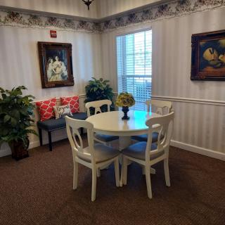 A cozy dining corner featuring a round table with four chairs, adorned with plants and paintings, next to a large window.