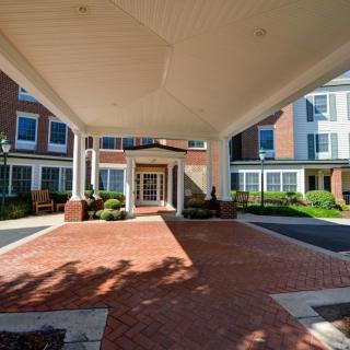 The front entrance of a building with a covered driveway and brick pathway, surrounded by well-maintained landscaping.