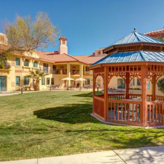 A sunny courtyard featuring a wooden gazebo and a surrounding building.