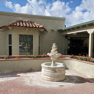 A small courtyard featuring a decorative fountain in front of a building with a tiled roof under a partly cloudy sky.