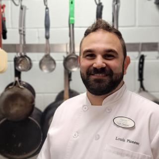 A chef smiling in a kitchen with cooking utensils hanging in the background.