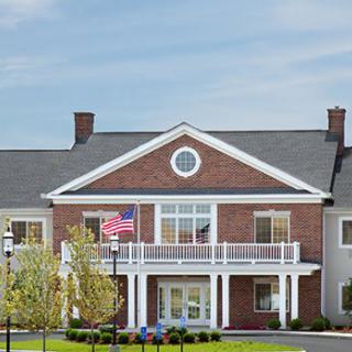 A two-story brick building with a white balcony and American flags displayed, surrounded by trees and a well-maintained lawn.