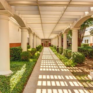 A sunlit corridor with columns casting shadows on a covered walkway surrounded by greenery.