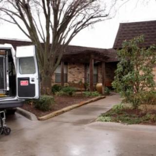 A van parked in the driveway of a brick house surrounded by trees and bushes on an overcast day.