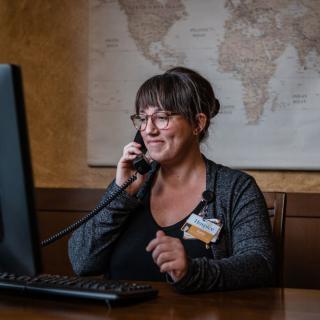 A woman in an office talking on the phone while sitting at a desk with a computer, with a map in the background.