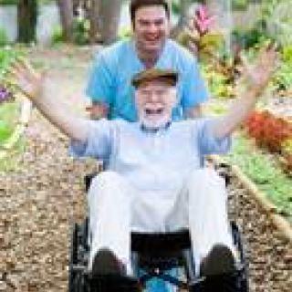 A joyful elderly man in a wheelchair being pushed by a smiling caregiver through a scenic garden path.
