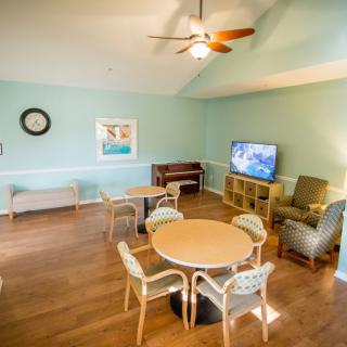 A cozy living room with wooden floors and light blue walls features a round table with chairs, a TV, sofas, and a ceiling fan.
