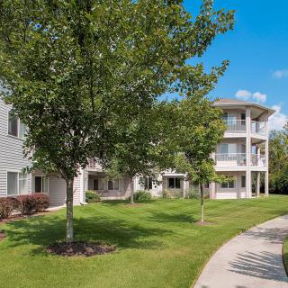 A residential apartment complex with landscaped lawns, trees, and a pathway on a sunny day.