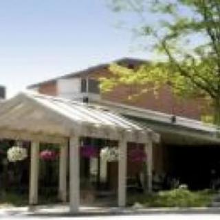 A building entrance with a glass canopy, surrounded by trees and flowers.