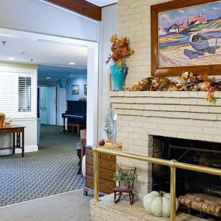 A cozy living room with a brick fireplace adorned with autumn decorations and a painting above, leading to an area with a piano.