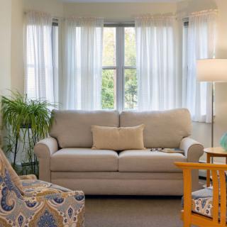 A cozy living room with a sofa, armchair, and floor lamp in front of a window with sheer curtains.