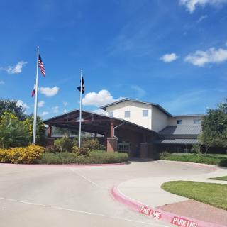 A welcoming entrance to a community building, featuring flags and well-maintained landscaping under a clear blue sky.