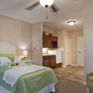 A cozy bedroom with a bed, side table, lamp, and a kitchenette area.