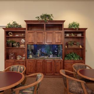 A cozy room with a large wooden cabinet housing an aquarium, surrounded by shelves decorated with plants and ornaments, with round tables and chairs nearby.
