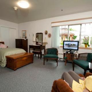 A cozy bedroom with a bed, dresser, chairs, and a television, warmly lit by natural light through the window.