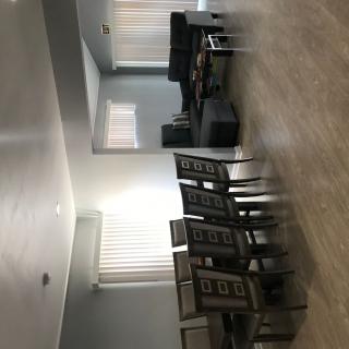 A spacious living room with a dining area, featuring a set of chairs and a table, complemented by large windows and hardwood flooring.